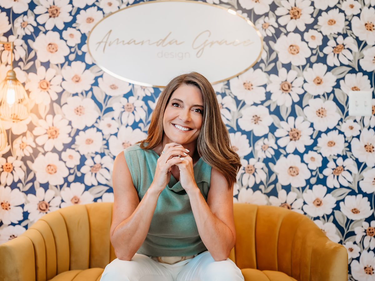 Mandy Corcoran smiling in a mustard chair in her studio, in front of floral wallpaper and an Amanda Grace Design sign